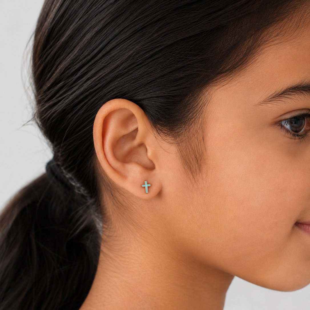 A close-up side profile of a young girl wearing a small silver cross stud earring on her earlobe, set against a clean light gray background.