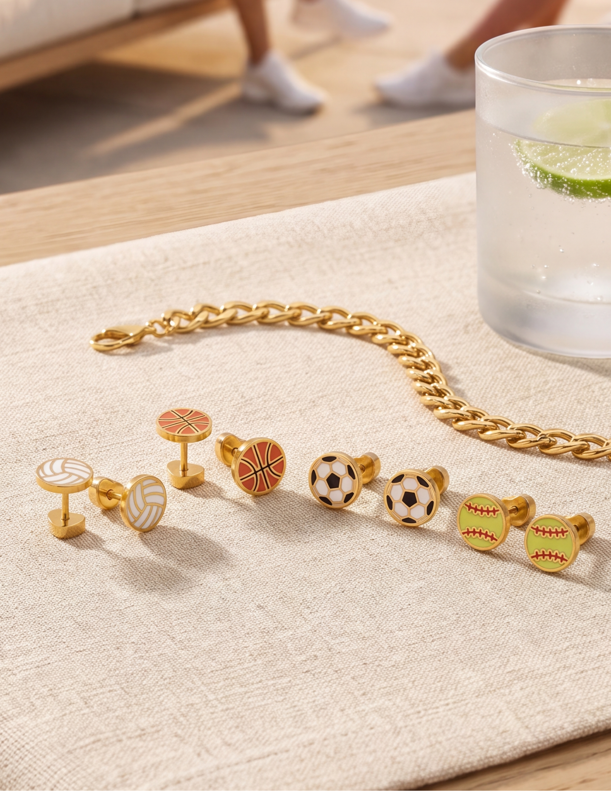 A set of gold-toned cufflinks with sports-themed enamel faces, including volleyball, basketball, soccer, football, baseball, and softball designs, arranged on a beige textured cloth next to a gold chain and a glass of water.