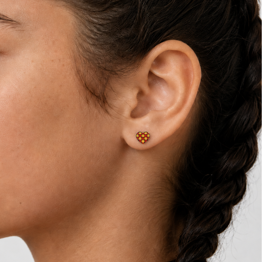 Close-up of a person wearing gold and red heart checkered stud earrings with a plain background