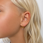 A close-up side profile of a young girl with blonde hair wearing a small gold hoop earring with a dangling purple cross charm, against a plain white background.