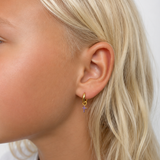 A close-up side profile of a young girl with blonde hair wearing a small gold hoop earring with a dangling purple cross charm, against a plain white background.