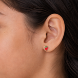 A close-up of a person's ear wearing a small strawberry-shaped stud earring with red enamel, green leaf accents, and a gold-tone outline.