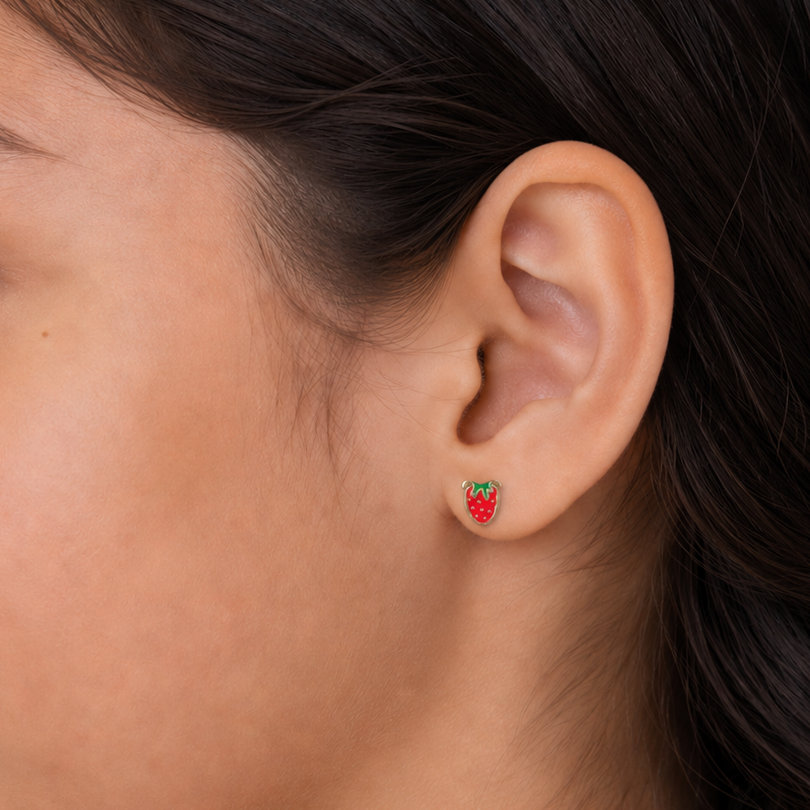 A close-up of a person's ear wearing a small strawberry-shaped stud earring with red enamel, green leaf accents, and a gold-tone outline.