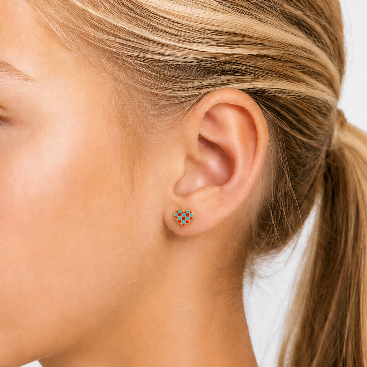 A close-up side profile of a young girl with blonde hair in a ponytail, wearing a small geometric stud earring with a blue and gold pattern against a clean white background.